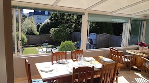 En solbeskinnet terrasse med et bord dækket til to, omgivet af stole, med udsigt over en have med træhegn og grønt.
