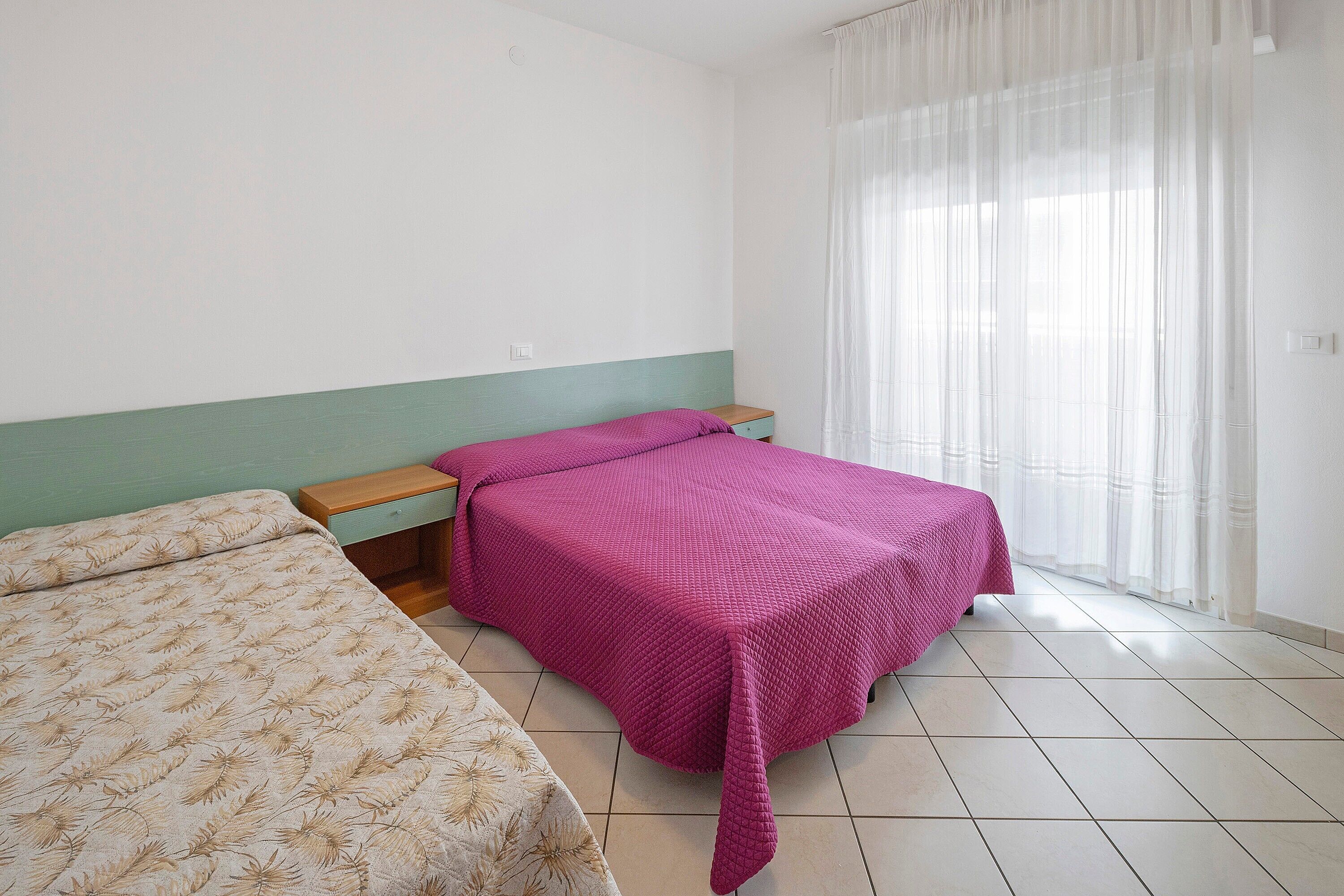 Camera da letto con due letti, un comodino in legno, una finestra con tende leggere e un pavimento piastrellato.