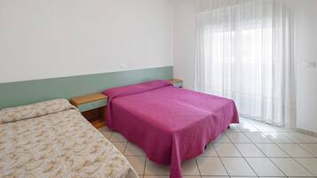 Camera da letto con due letti, un comodino in legno, una finestra con tende leggere e un pavimento piastrellato.