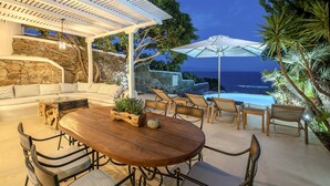 An outdoor patio with a stone wall, a wooden table with chairs, a sofa, and an umbrella. - Villa Tiche with private pool - My Rental Homes (Mykonos)