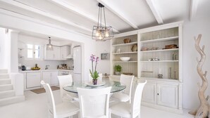 A modern kitchen with white cabinetry, a glass dining table, and white chairs. There is a built-in bookshelf with decorative items and a wooden sculpture. - Villa Tiche with private pool - My Rental Homes (Mykonos)