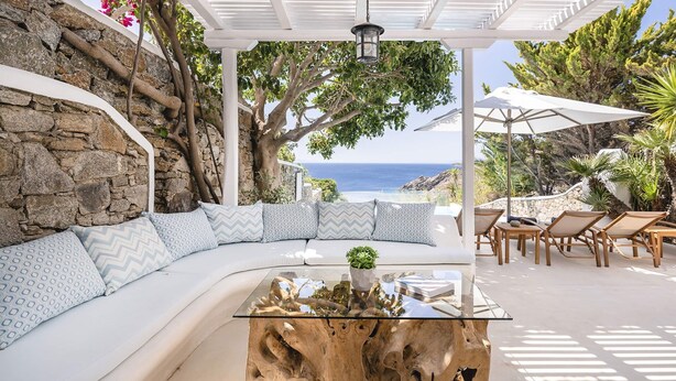 A modern outdoor seating area with a curved white sofa, a glass coffee table, and a view of the sea. - Villa Tiche with private pool - My Rental Homes (Mykonos)