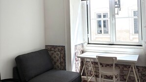 A room with a sofa, a small table, a chair, a window with a view of buildings, and patterned wall tiles. - SJ1D Warm & Inviting | Historic Stay! | A/C (Lisboa ciudad)