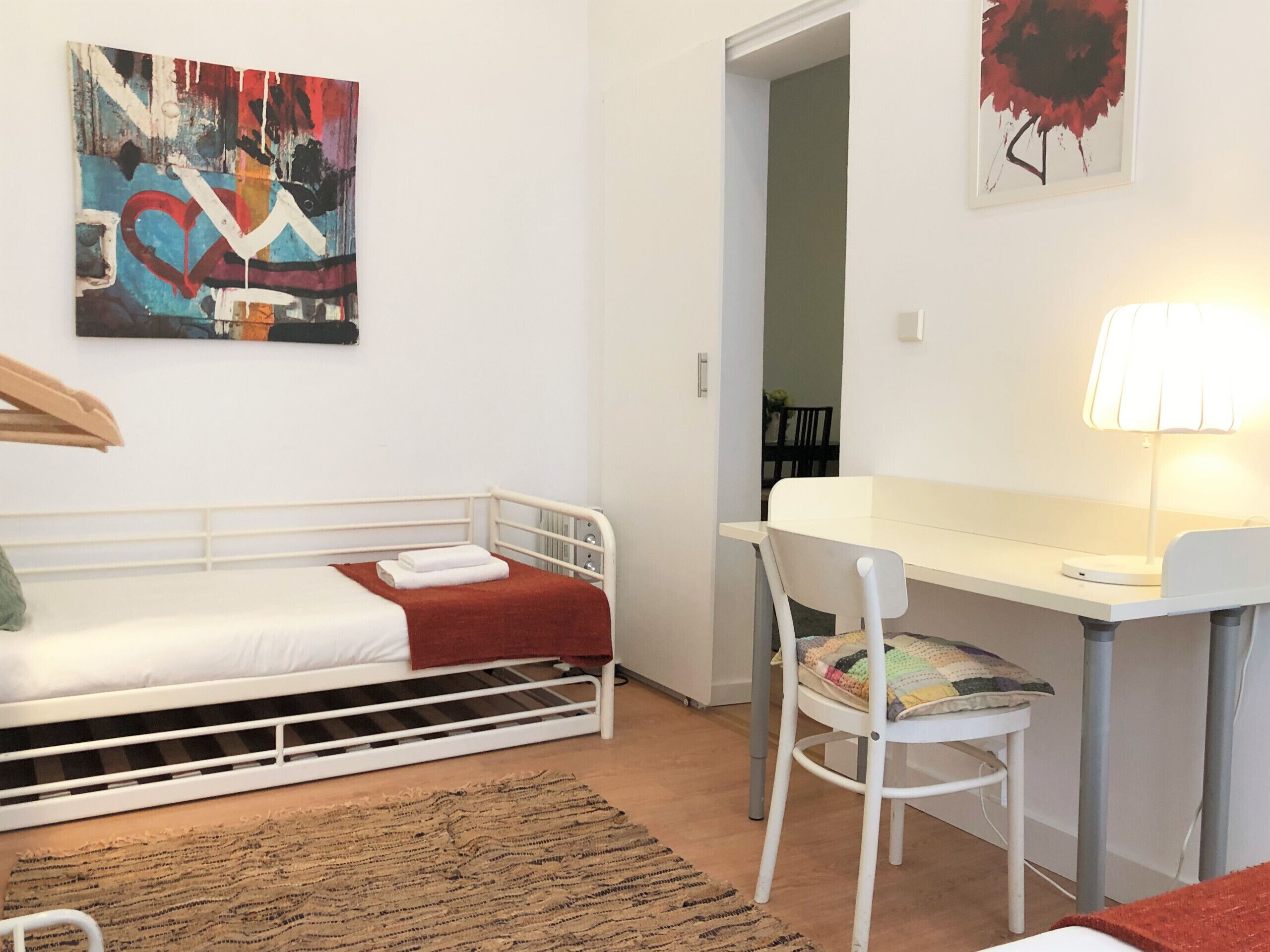 A small, modern room with a single bed, a desk, and a chair. There is a painting on the wall and a lamp on the desk.