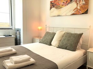 A neatly made bed with white and grey linens, a bedside lamp, and a painting above it. - SJ1E Spacious & Cozy | Downtown - Baixa | A/C (Lisboa ciudad)