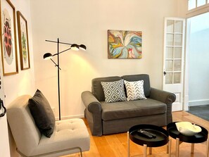 Ein Wohnzimmer mit grauem Sofa, einem Sessel, zwei schwarzen Beistelltischen, einer Stehlampe und einem farbenfrohen, abstrakten Gemälde an der Wand.