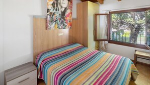 A bedroom with a bed, a nightstand, a window with a view of trees, and a colorful painting on the wall. - Apartment 'Borgo Stella X' with Private Terrace, Wi-Fi and Air Conditioning (Bibione)