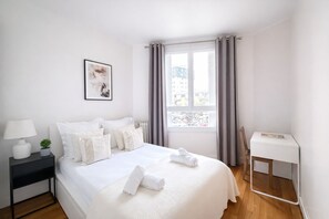 Habitación de hotel ordenada con una cama grande, una mesita de noche con lámpara, una planta pequeña y una ventana con cortinas.