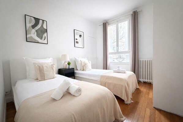 Una habitación de hotel con dos camas, una mesita de noche con lámpara, una ventana con cortinas y un radiador.