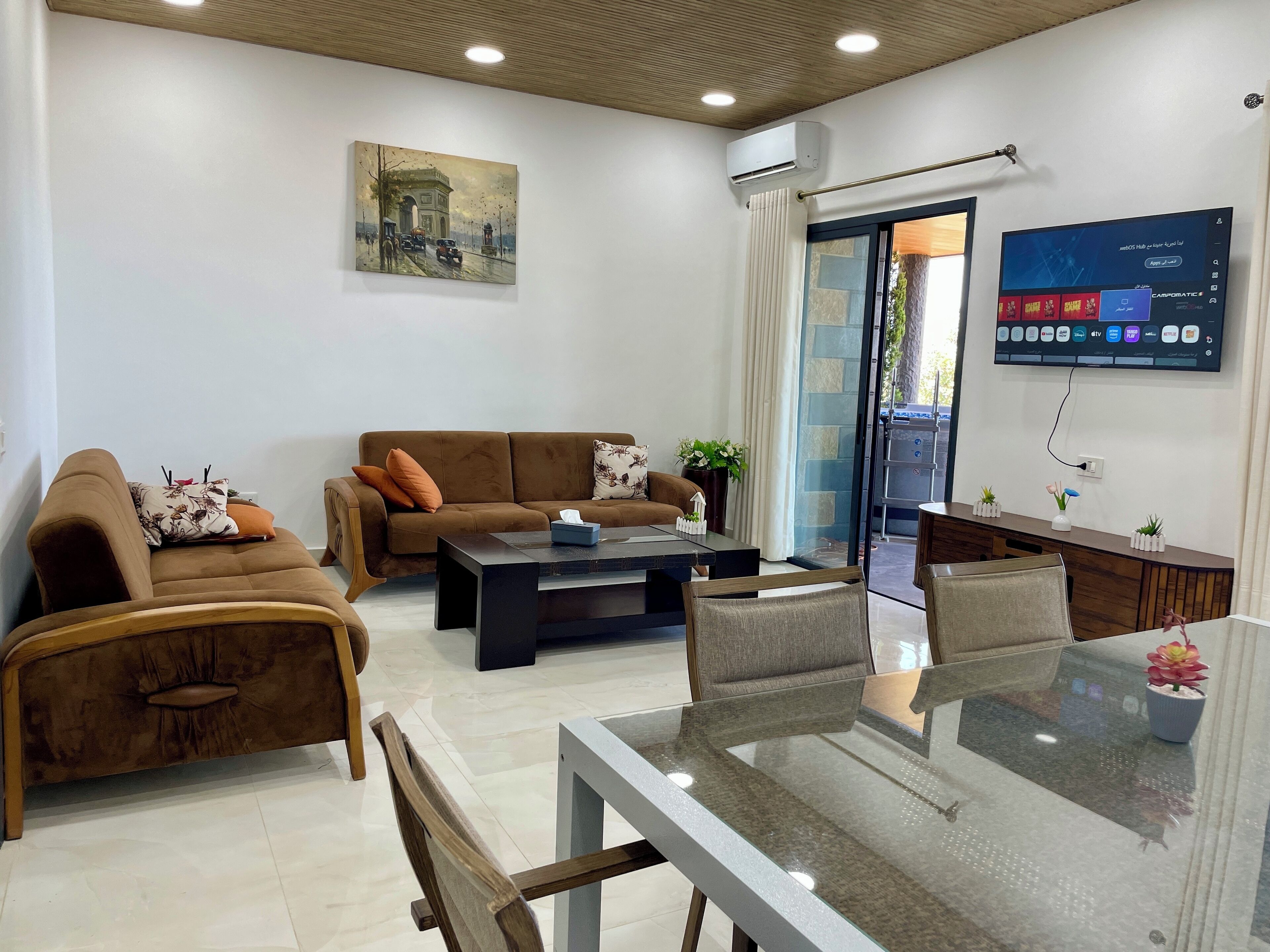 A modern living room with a brown sofa, a glass coffee table, and a TV mounted on the wall.