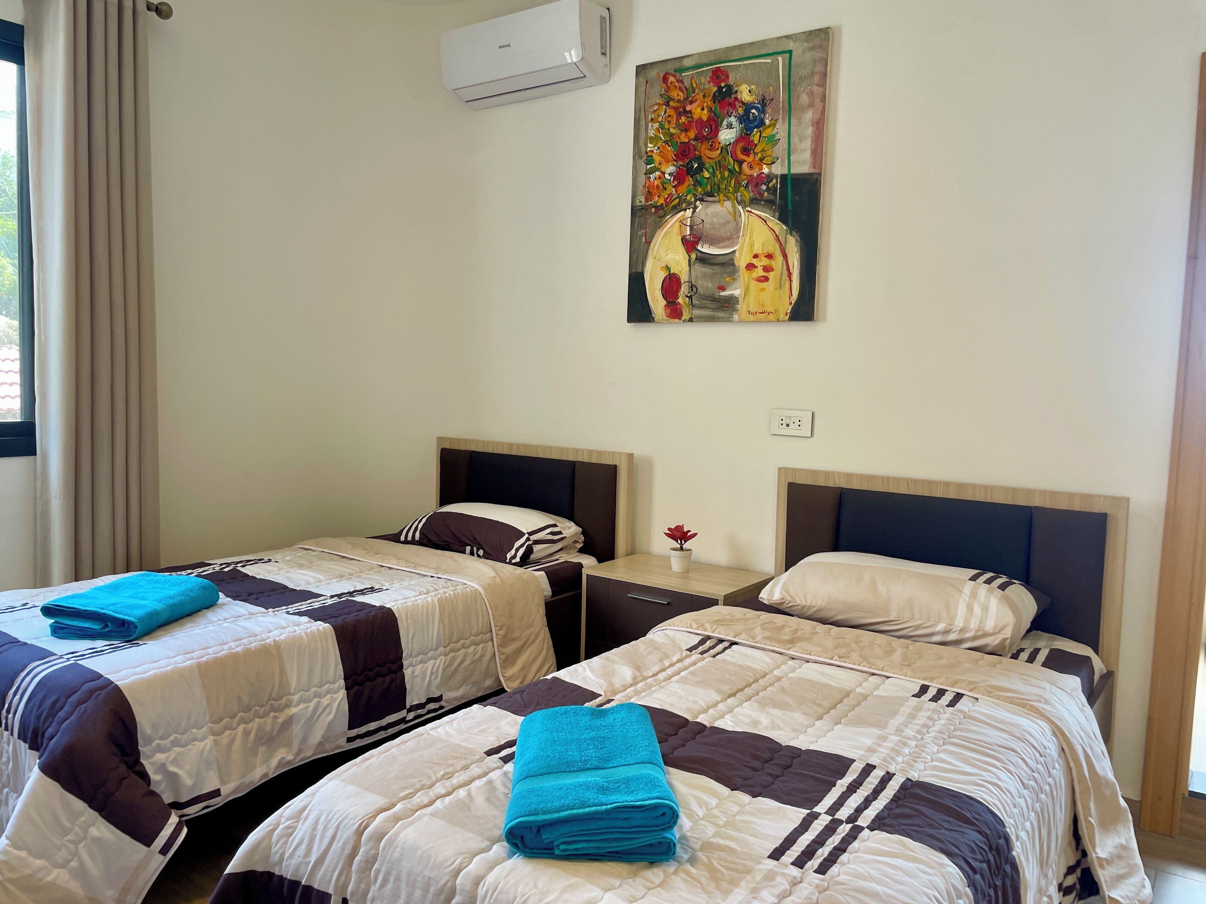 A hotel room with two beds, each with a different patterned bedspread and pillows, a small table with a potted plant, and a painting of a vase with flowers on the wall.