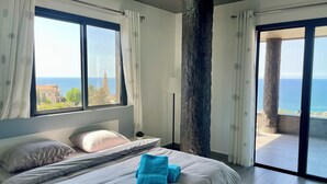 A bedroom with a bed, pillows, a blue towel, and a view of the sea through large windows. - Kfaraabida SKY (Batroun)