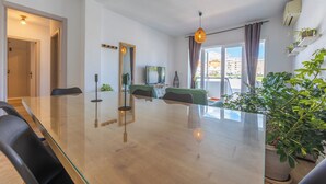 Een moderne woonkamer met een glazen eettafel, comfortabele zitplaatsen, een televisie en een balkon met planten.