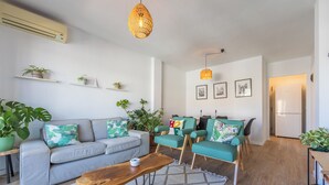 Een moderne woonkamer met een grijze bank, blauwgroene fauteuils en een houten salontafel. Er staan potplanten, een hanglamp en er hangen foto's aan de muur.