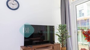 A modern living room with a flat-screen TV on a wooden TV stand, a wall clock, and a potted plant. - Bloomfields Iconic 1BR in Oasis Masd (Abu Dhabi)