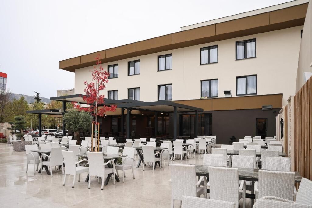 An outdoor patio area with white wicker chairs and tables, a covered seating area, and a building with multiple windows and a flat roof.