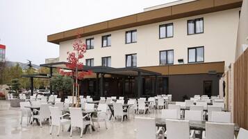 An outdoor patio area with white wicker chairs and tables, a covered seating area, and a building with multiple windows and a flat roof.