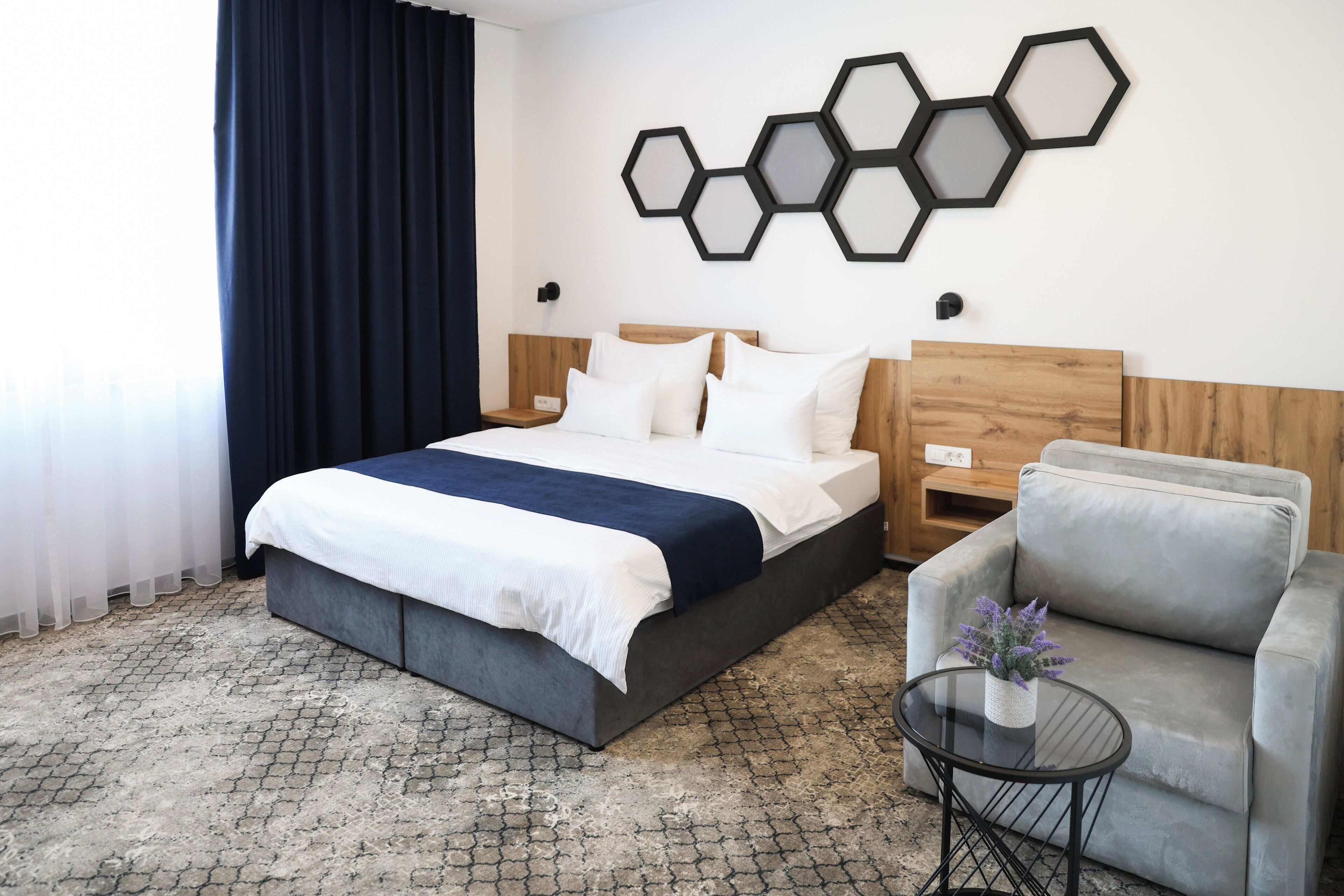 A hotel room with a bed, a grey sofa, a small round table with a plant, and a hexagonal wall decoration.