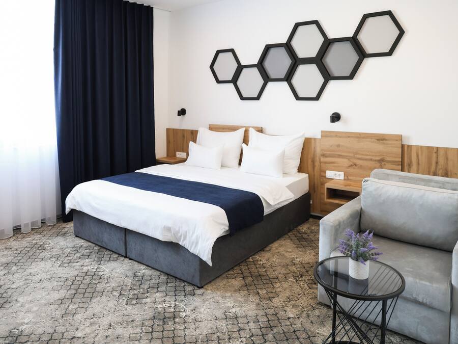 Habitación de hotel con cama, sofá gris, mesita redonda con planta y decoración hexagonal en la pared.