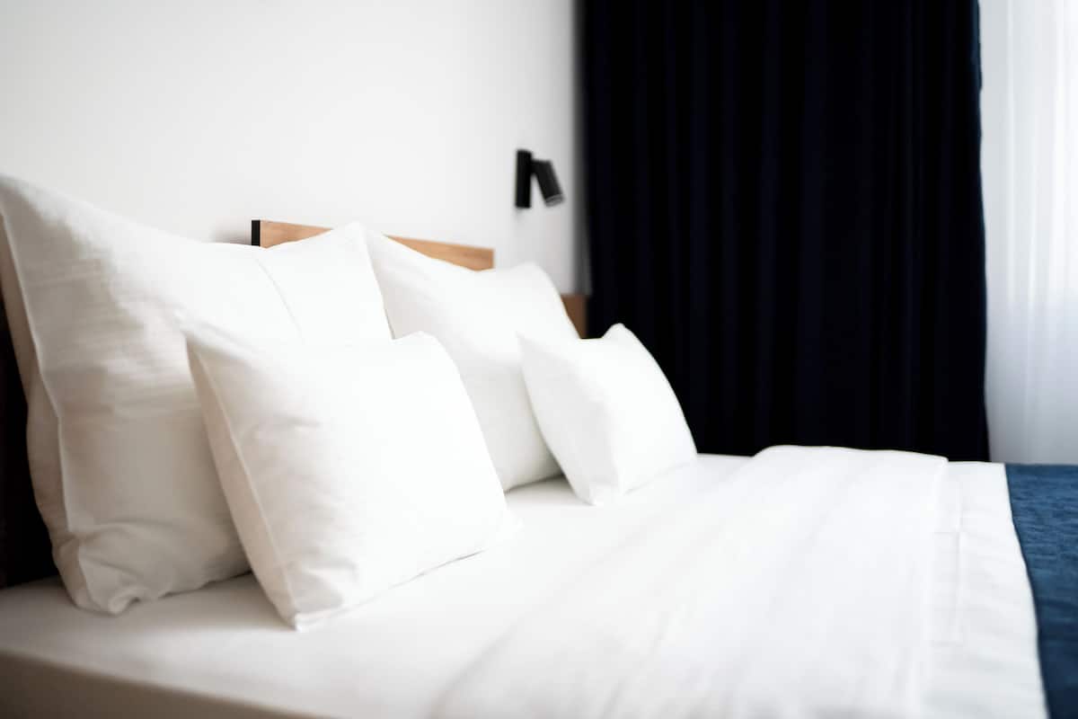 A neatly made bed with white bedding and pillows, a wooden headboard, and a dark curtain partially drawn to the side.