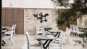 An outdoor seating area with white wicker chairs and tables, a stone wall with a black abstract art piece, and a wooden fence in the background. - Hotel Karting (Mostar)