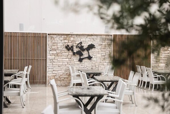 An outdoor seating area with white wicker chairs and tables, a stone wall with a black abstract art piece, and a wooden fence in the background. - Hotel Karting (Mostar)