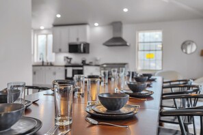 A dining table set with plates, bowls, glasses, and cutlery in a modern kitchen with white cabinets and a large window.