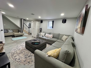 A living room with a sectional sofa, a coffee table, a patterned rug, a staircase, and a painting on the wall. - World Cup Special: Spacious home in secluded woods close to events and NYC. (Watchung)
