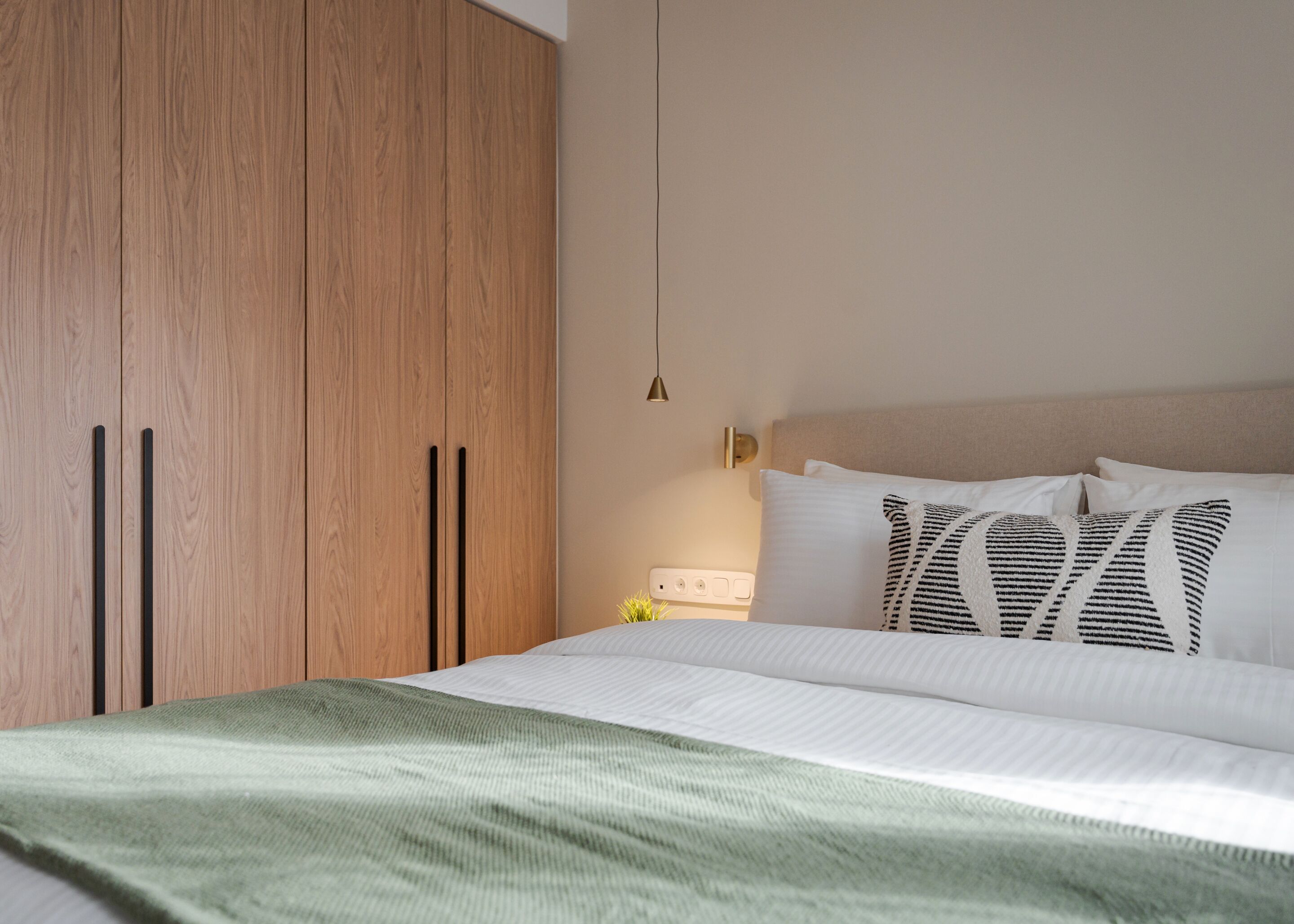 Quarto com cama, manta verde, guarda-roupa de madeira e mesa de cabeceira com abajur.