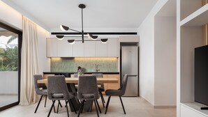 Un comedor moderno con mesa de madera y sillas grises tapizadas, una cocina integrada con refrigerador de acero inoxidable y un amplio ventanal con cortinas.