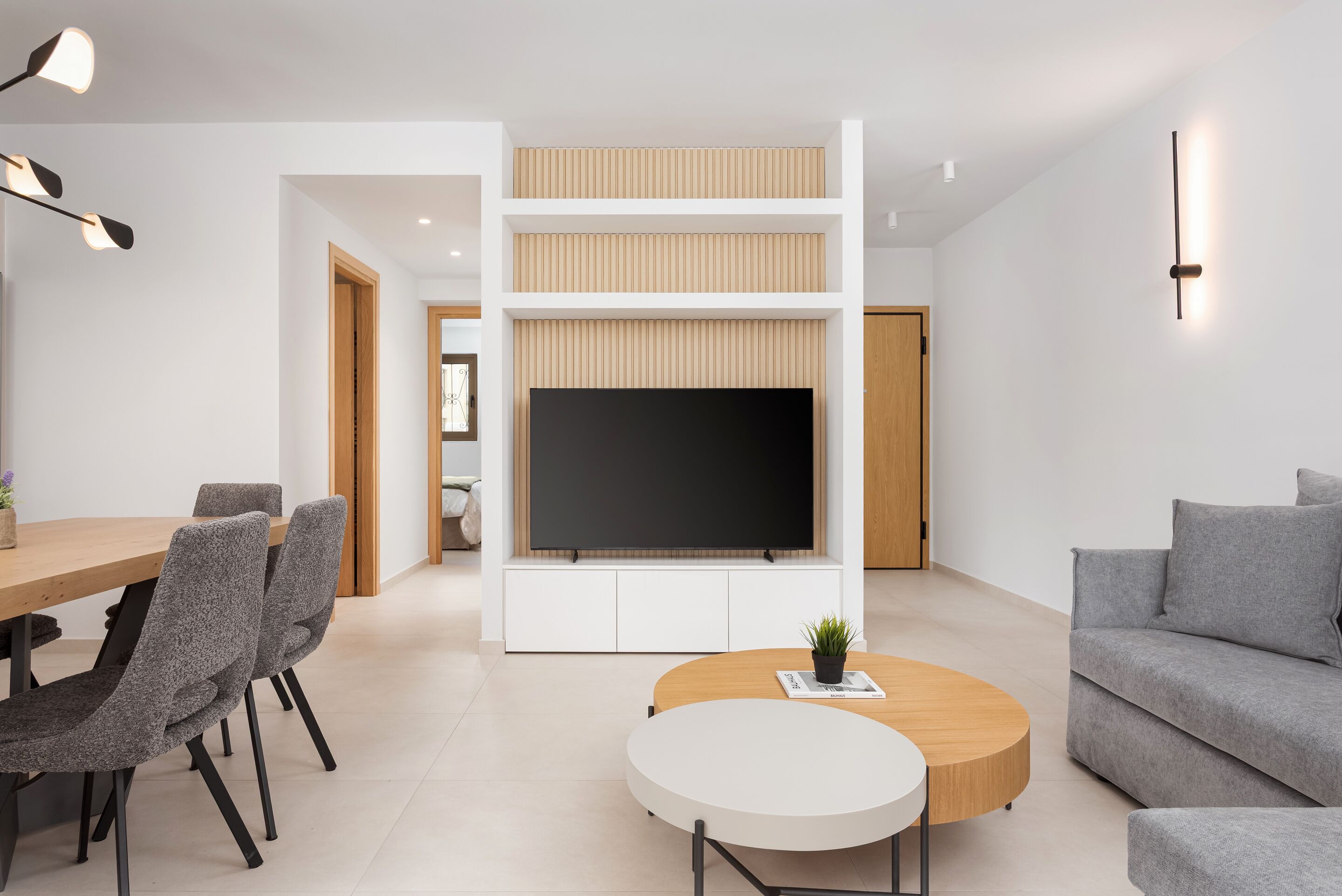 A modern living room with a flat-screen TV, a wooden coffee table, grey sofas, and a dining area with a wooden table and grey chairs.