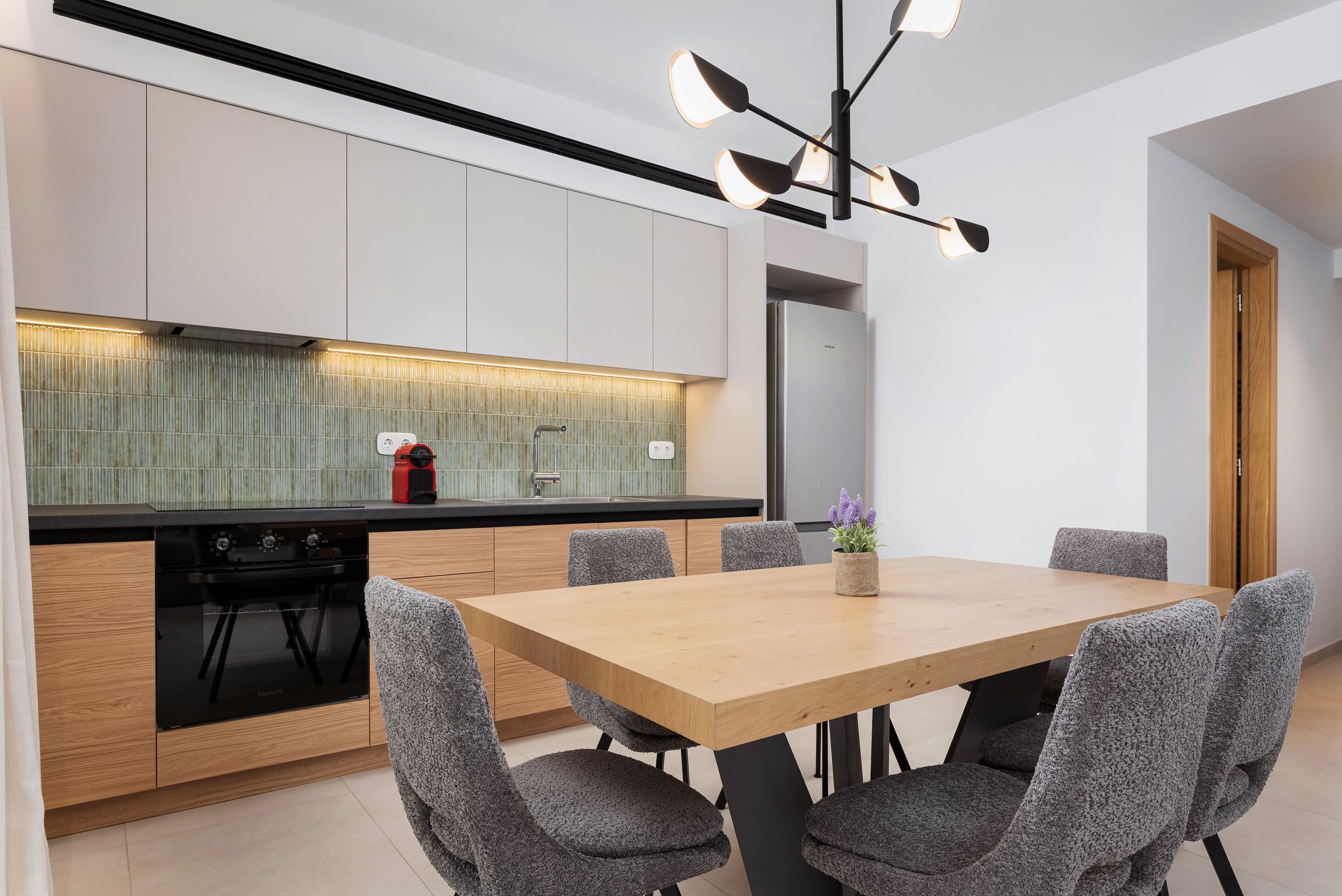 A modern kitchen with a wooden table and grey upholstered chairs, a built-in oven, and a stainless steel refrigerator.