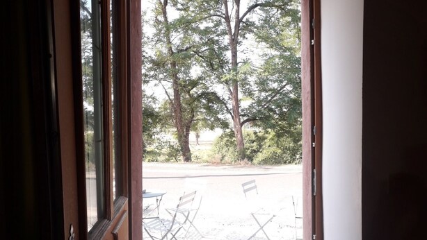 An open door leading to an outdoor seating area with a gravel ground, a bench, and trees in the background.
