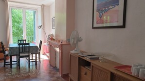 Una habitación con una mesa de comedor, sillas, un radiador, un ventilador, una cubierta para radiador, un cuadro en la pared y una ventana con cortinas.