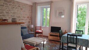 Una sala de estar con un sofá, un televisor sobre un mueble de madera, una mesa de comedor con sillas, un tapete estampado y una ventana con cortinas.