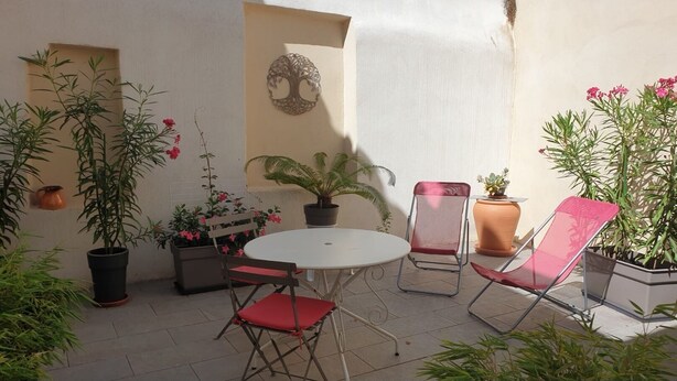 Un patio con una mesa redonda y dos sillas, rodeado de plantas en macetas y un adorno de árbol sujeto a la pared.