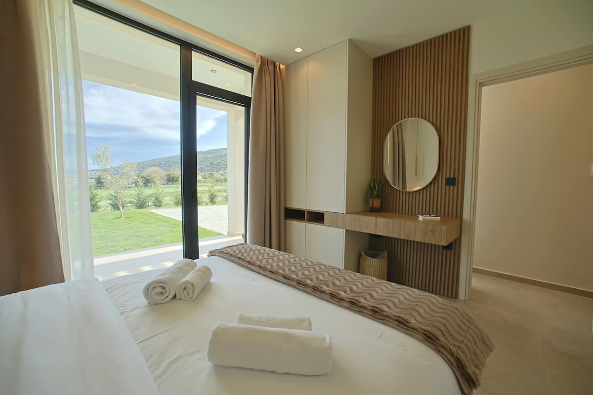 A modern hotel room with a large bed, a view of the outdoors, a built-in vanity, and a large mirror.