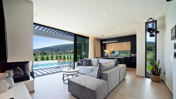 A modern living room with a sectional sofa, a coffee table, and a view of a pool and hills. - Ioannina Secret Luxury Villas (Vounoplagia)