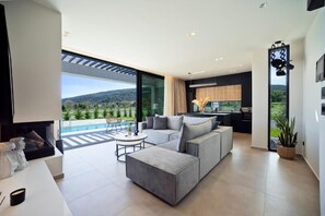 A modern living room with a sectional sofa, a coffee table, and a view of a pool and hills. - Ioannina Secret Luxury Villas (Vounoplagia)