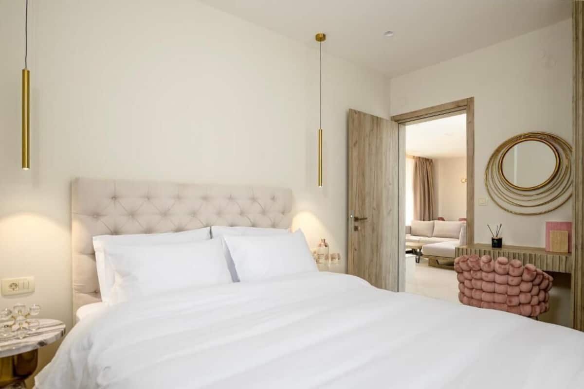 A bedroom with a tufted headboard, a large bed, bedside tables, a mirror, and a door leading to another room.