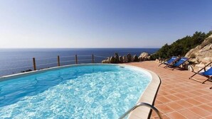 Una piscina con vista despejada al océano, rodeada por una terraza de baldosas y sillas reclinables azules.