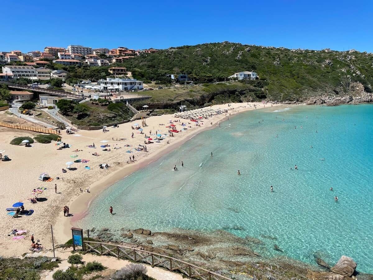A coastal beach with clear turquoise waters, a sandy shore, and buildings in the background.