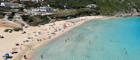 A coastal beach with clear turquoise waters, a sandy shore, and buildings in the background.
