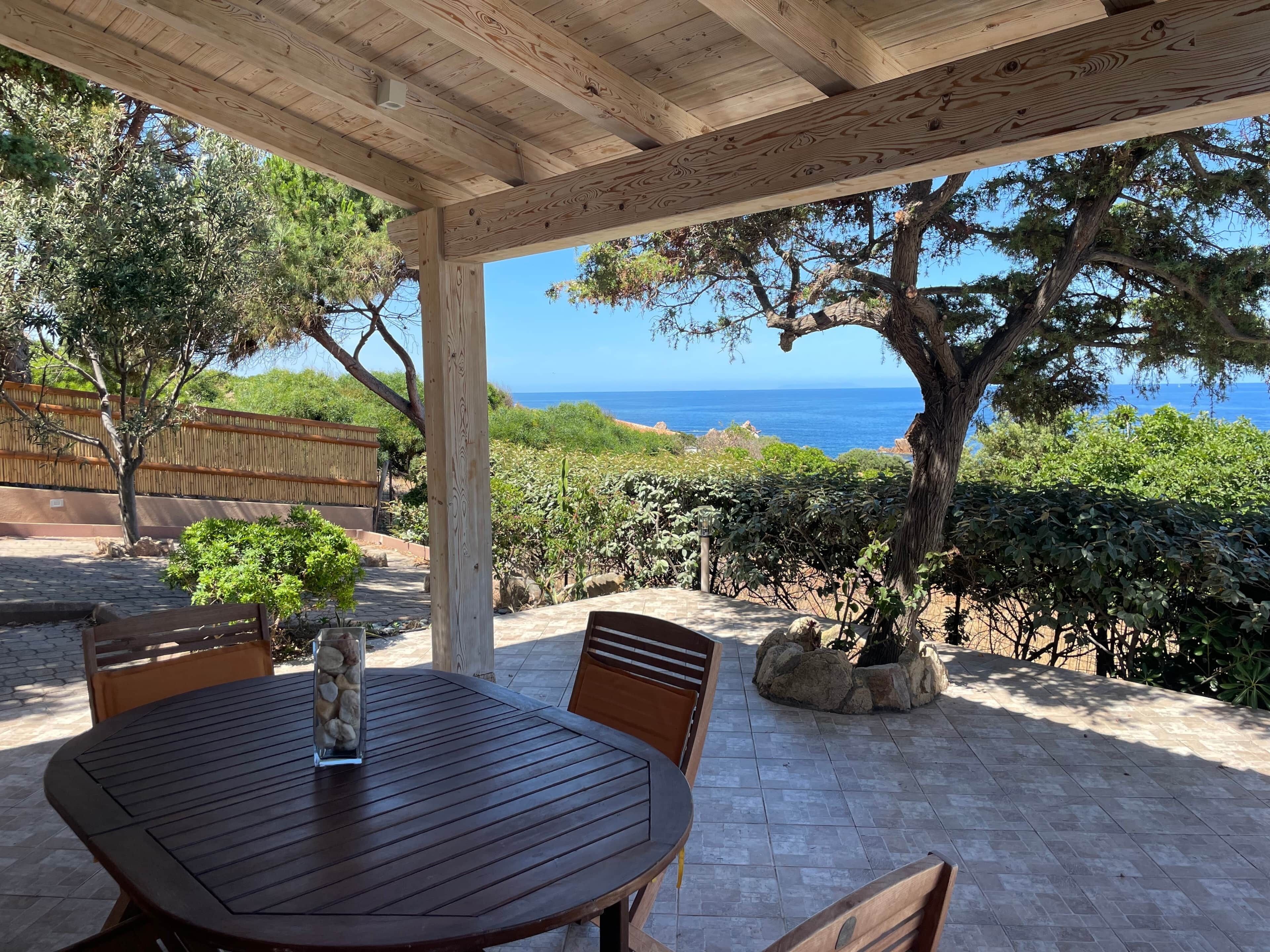 Een houten pergola met een tafel en stoelen, met uitzicht op een kustlandschap met bomen en zee.