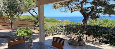 A wooden pergola with a table and chairs, overlooking a coastal landscape with trees and a view of the sea.
