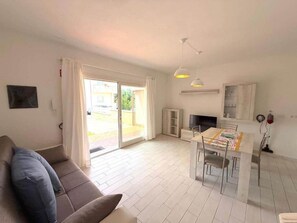 A living room with a sofa, a dining table with chairs, a TV, and a sliding glass door leading to an outdoor area. - Villetta Acacia K&G House (Viddalba)