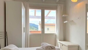 Una camera da letto con un letto, un comodino, una sedia e una finestra vista edifici e colline.