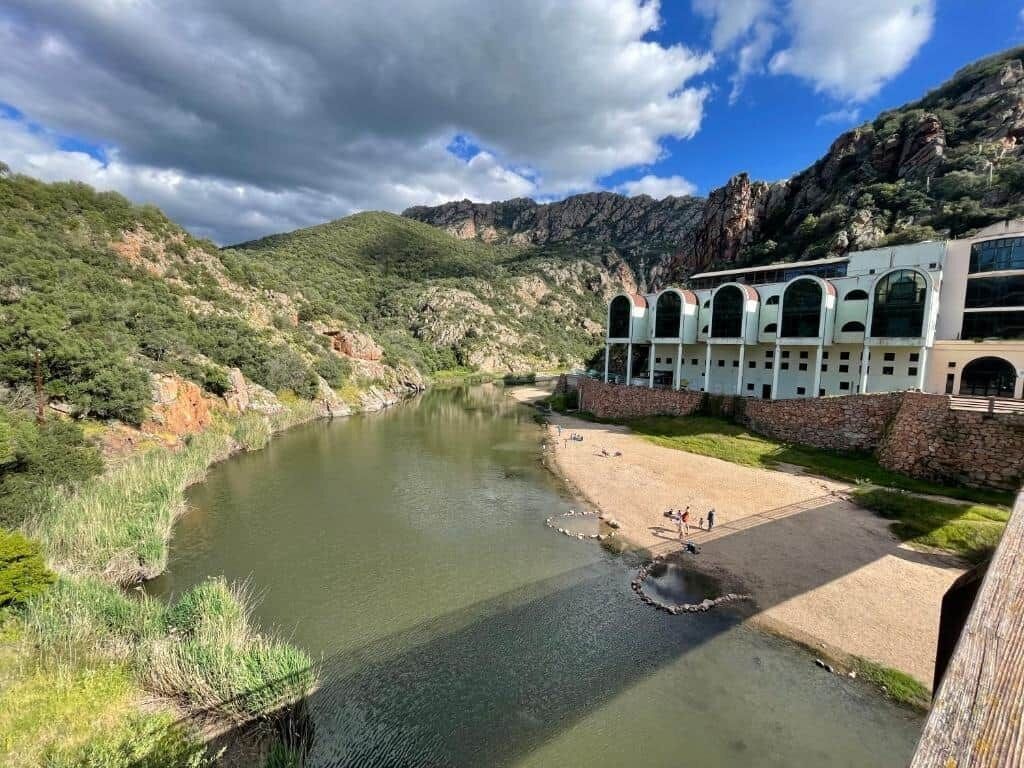 A river with a bridge, a building with arched windows, and a mountainous backdrop.