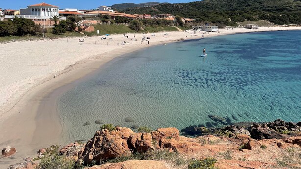 Una playa costera con aguas cristalinas, orilla arenosa y edificios al fondo.