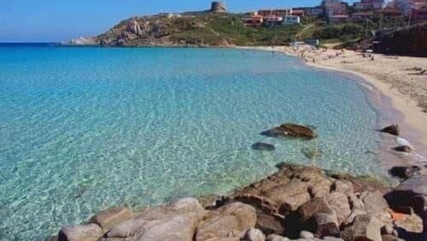 Una playa costera con agua turquesa cristalina, costa rocosa y edificios al fondo.